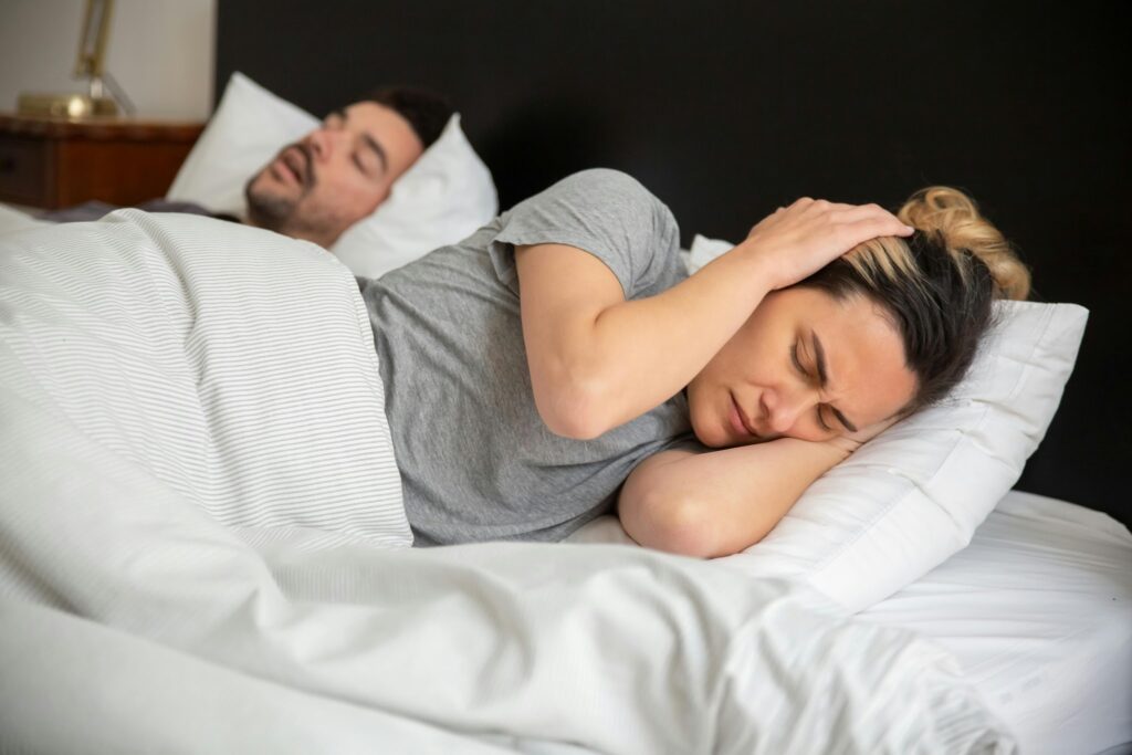 Woman covering ears while man shows signs of sleep apnea and snoring. He has obstructive sleep apnea symptoms. The woman worries and wonders: “Can sleep apnea kill you?” She wants to get him an obstructive sleep apnea test.