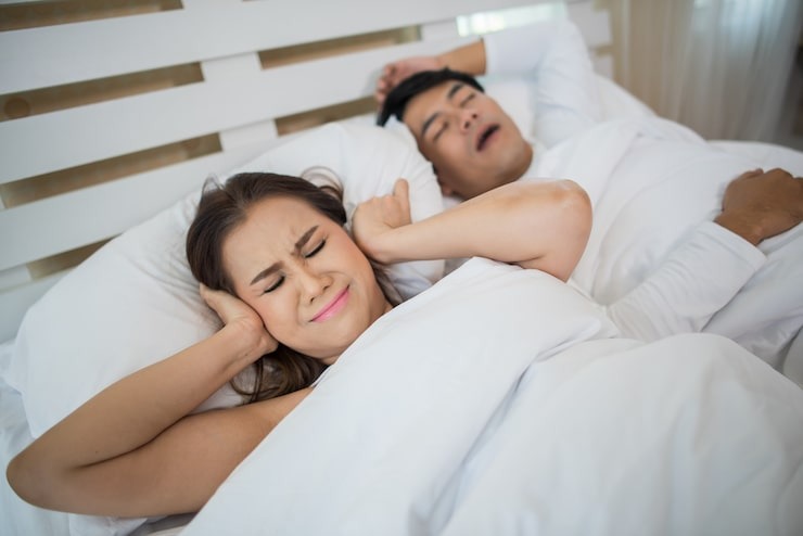 Woman covering her ears in frustration while her partner snores loudly in bed, illustrating sleep disturbances from sleep apnea.